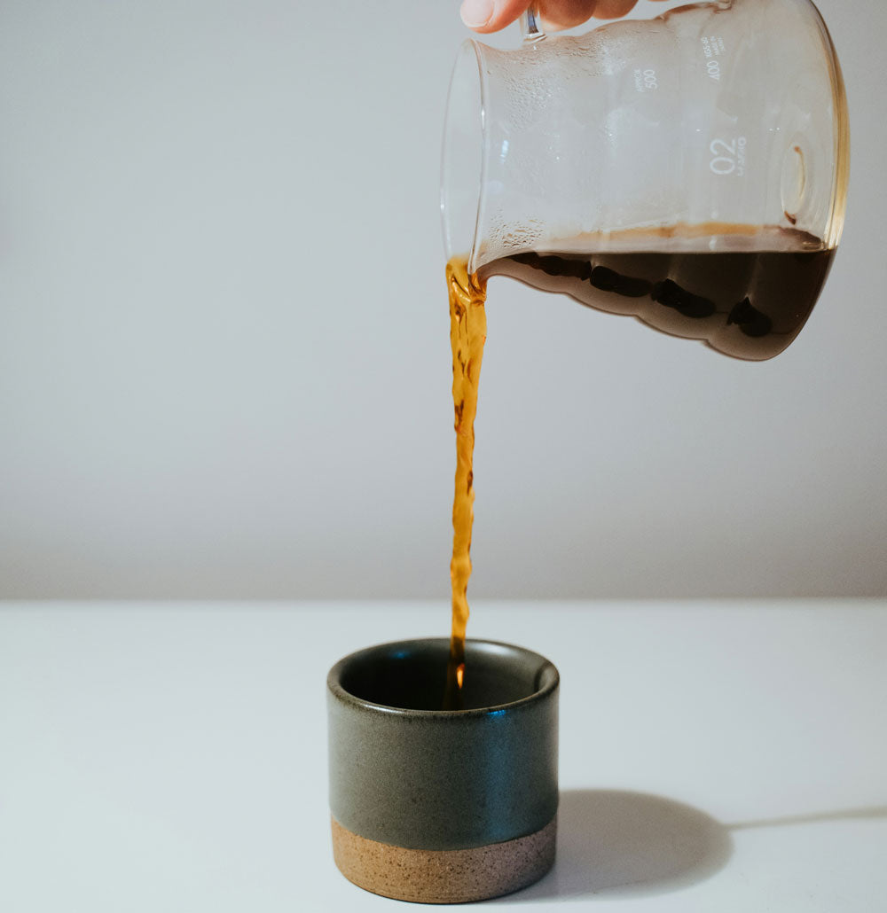 Coffee being poured from a glass carafe into a ceramic cup on a white surface.